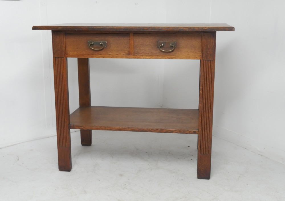 An Arts and Crafts solid oak side table featuring two drawers with brass handles, fluted legs, and a lower shelf.