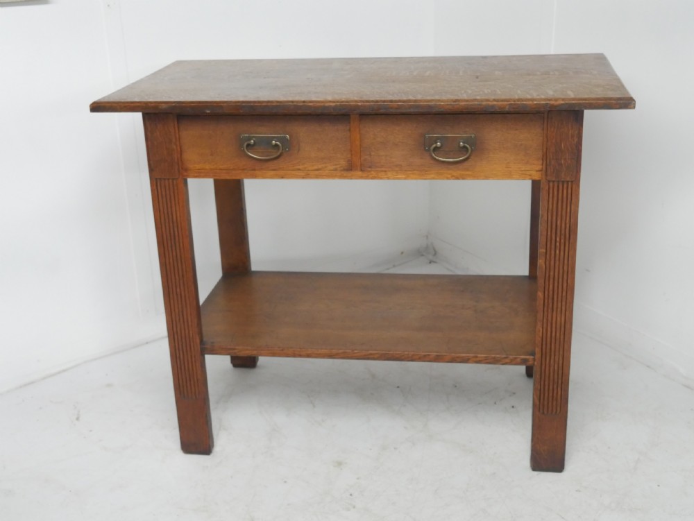 An Arts and Crafts solid oak side table featuring two drawers with brass handles, fluted legs, and a lower shelf. View II