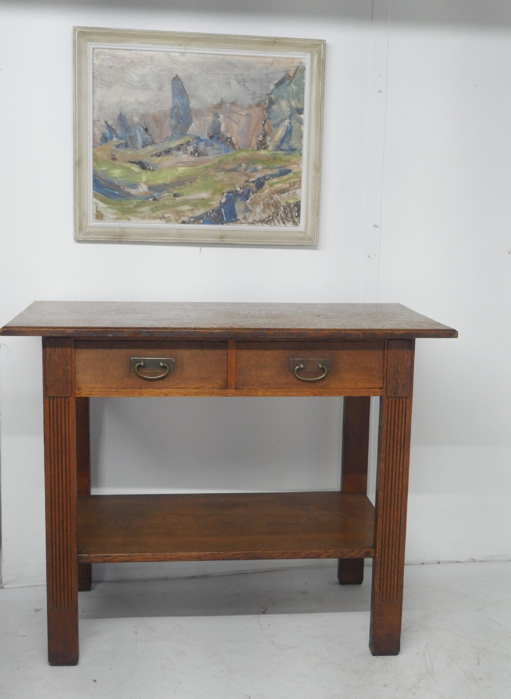 An Arts and Crafts solid oak side table featuring two drawers with brass handles, fluted legs, and a lower shelf. View X