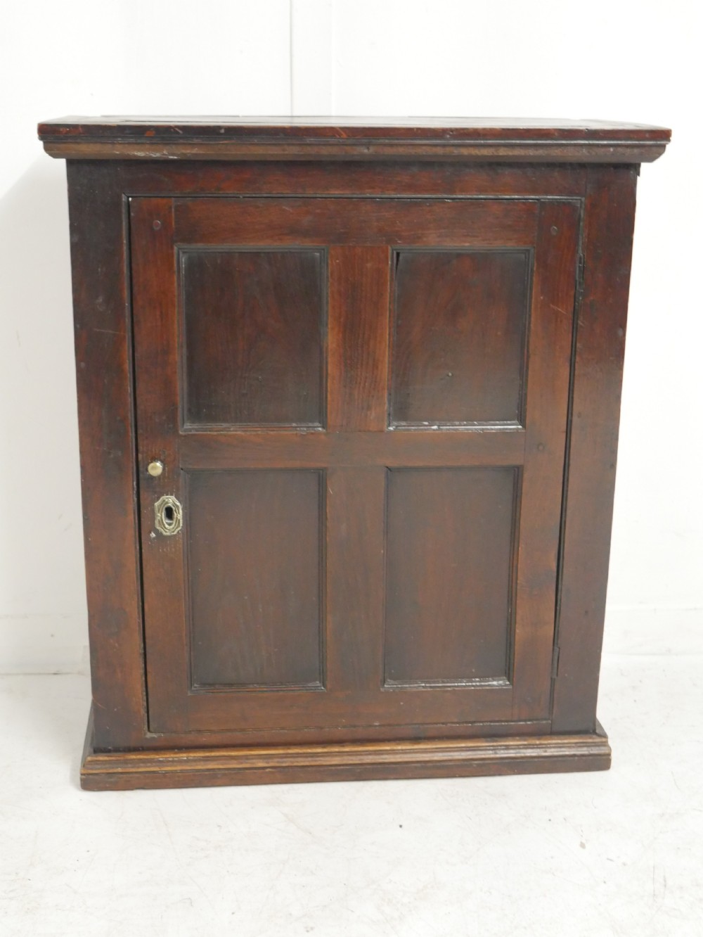 A dark brown Georgian oak wall cupboard with a single four-panel door, brass keyhole escutcheon, and moulded top cornice.