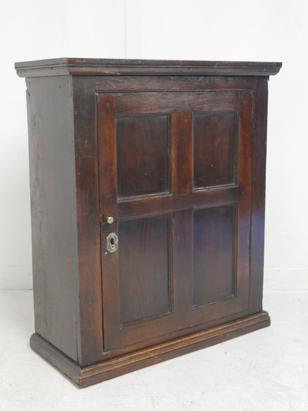 A dark brown Georgian oak wall cupboard with a single four-panel door, brass keyhole escutcheon, and moulded top cornice. View III