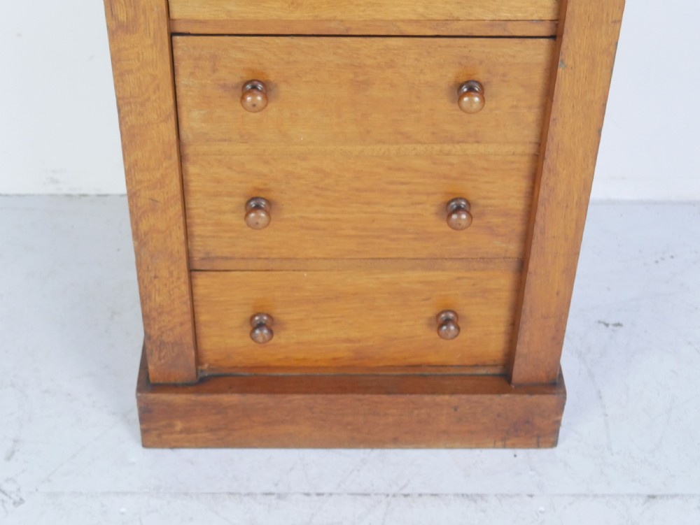 A tall Victorian oak Wellington chest featuring seven drawers with turned wooden knob handles, carved corbels at the top corners, and a locking side flap. View III