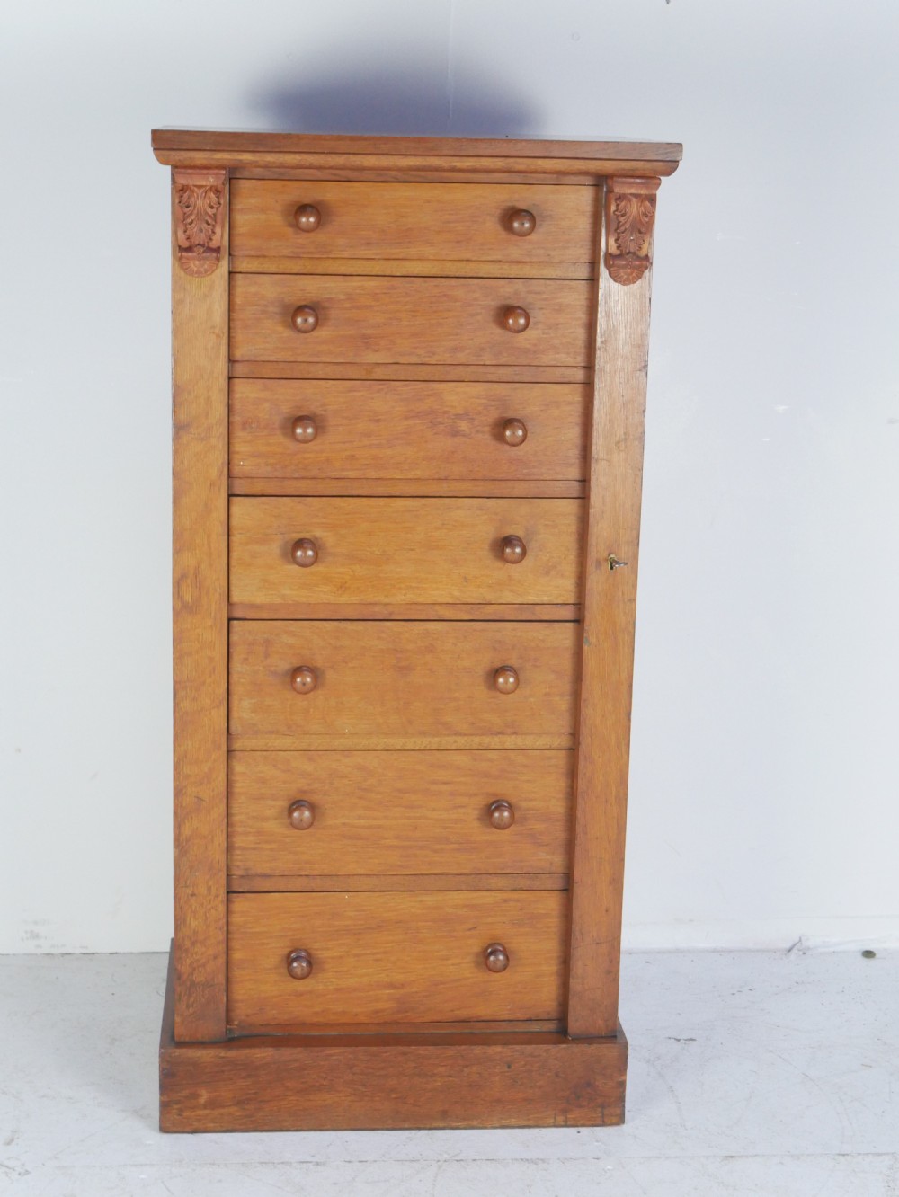 A tall Victorian oak Wellington chest featuring seven drawers with turned wooden knob handles, carved corbels at the top corners, and a locking side flap.