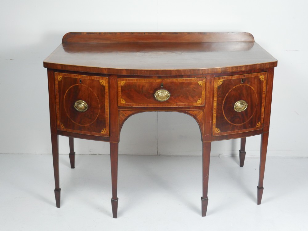 A Georgian bow-front mahogany sideboard featuring satinwood inlays, brass oval handles, and square tapered legs. View XVI