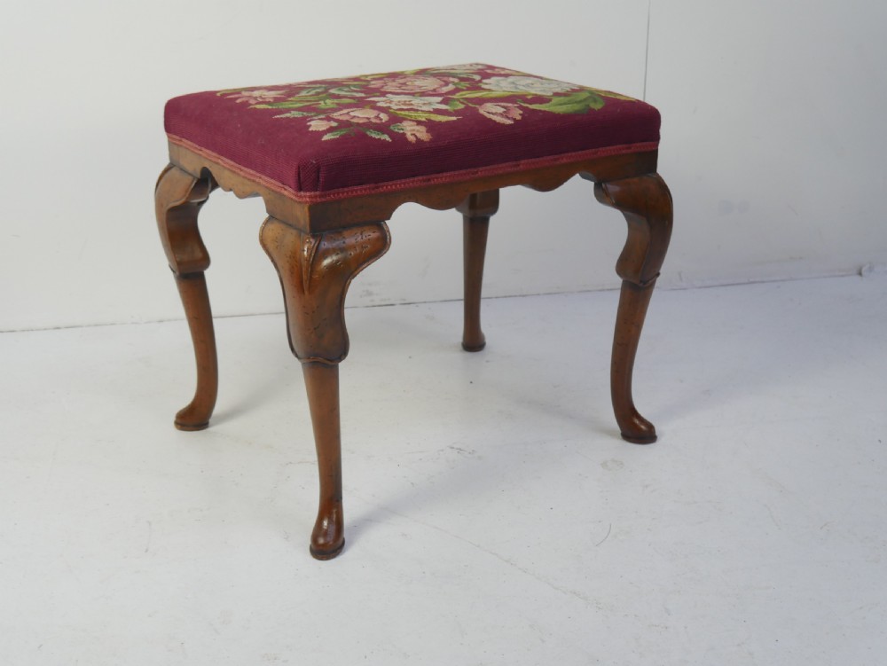 A 1920s low wooden footstool featuring carved cabriole legs and a rectangular seat upholstered in burgundy floral needlepoint tapestry.
