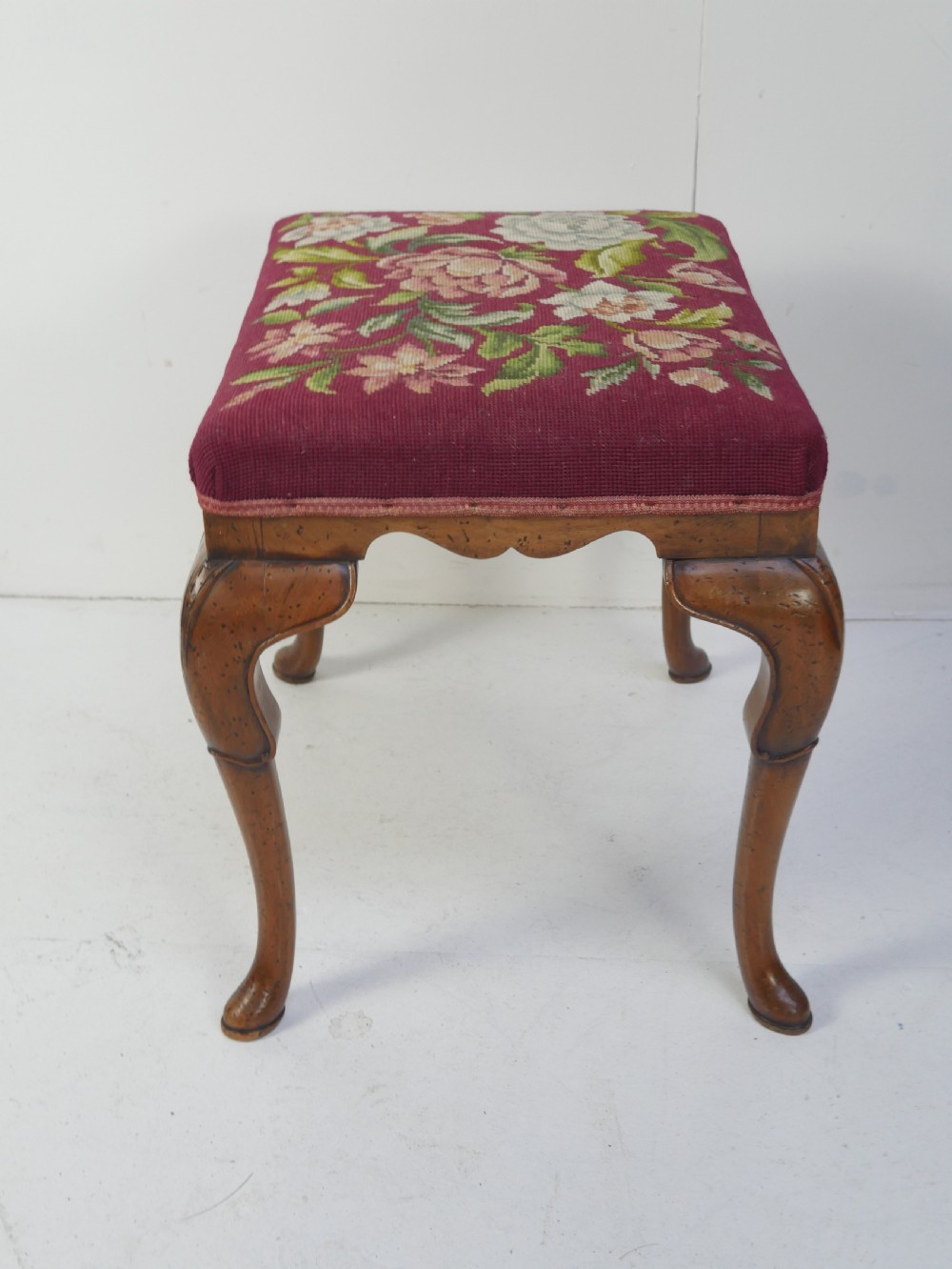 A 1920s low wooden footstool featuring carved cabriole legs and a rectangular seat upholstered in burgundy floral needlepoint tapestry. View VII