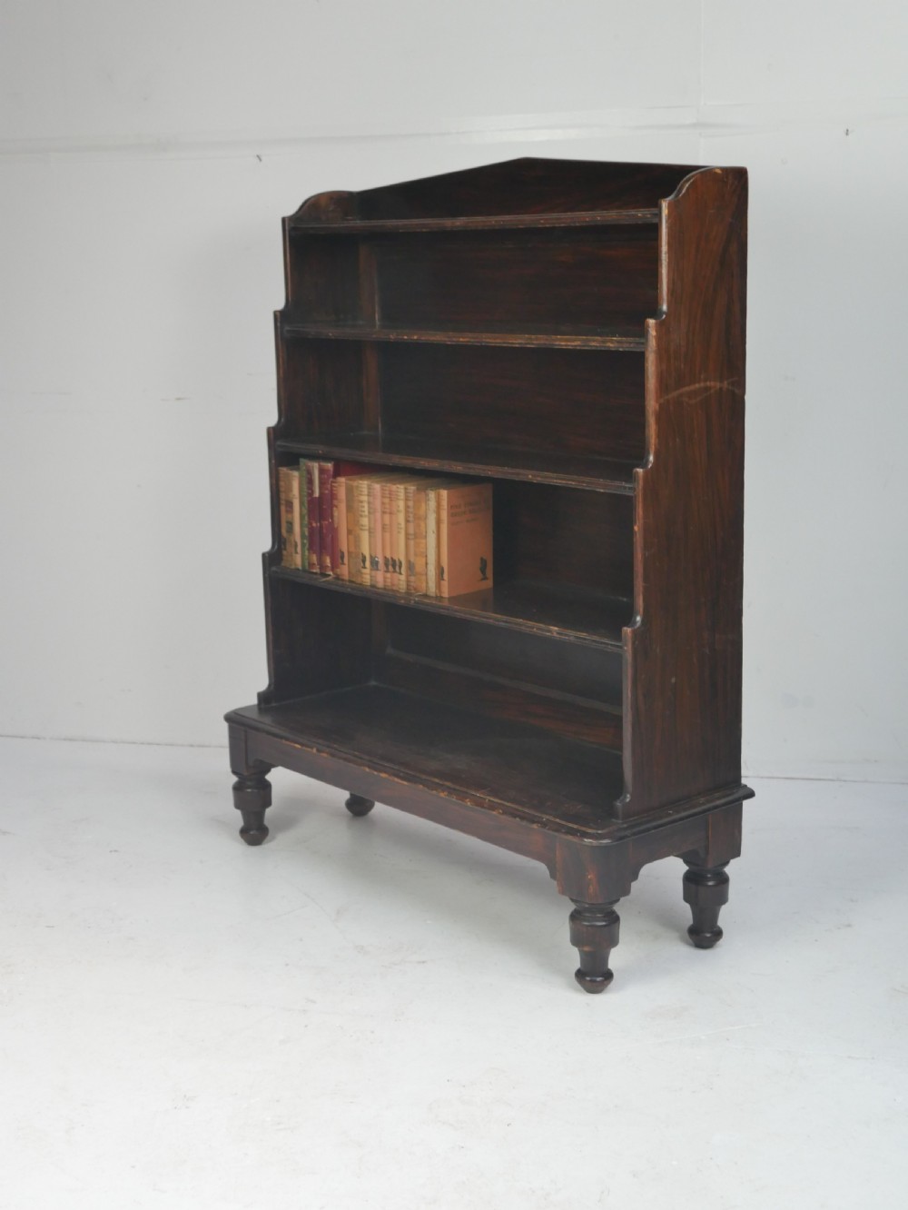 A dark brown 19th-century waterfall bookcase featuring four open stepped shelves, side profile cutouts, and turned legs.