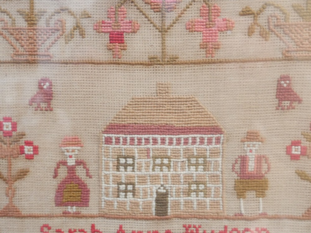 An 1856 Victorian needlework sampler featuring embroidered house, figure, bird, and floral motifs inside a golden maple wood frame. View VII
