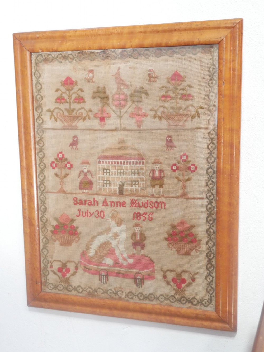An 1856 Victorian needlework sampler featuring embroidered house, figure, bird, and floral motifs inside a golden maple wood frame.