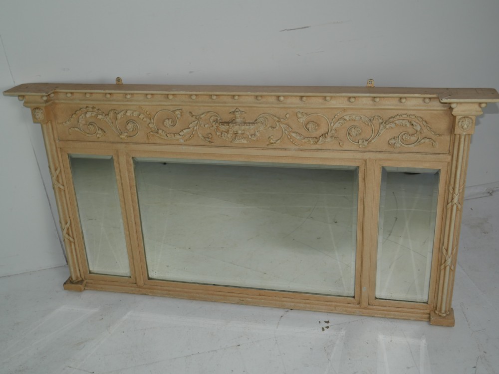 A wide cream-painted neoclassical overmantel mirror featuring three glass panels, an ornate carved urn frieze, and side pilasters.