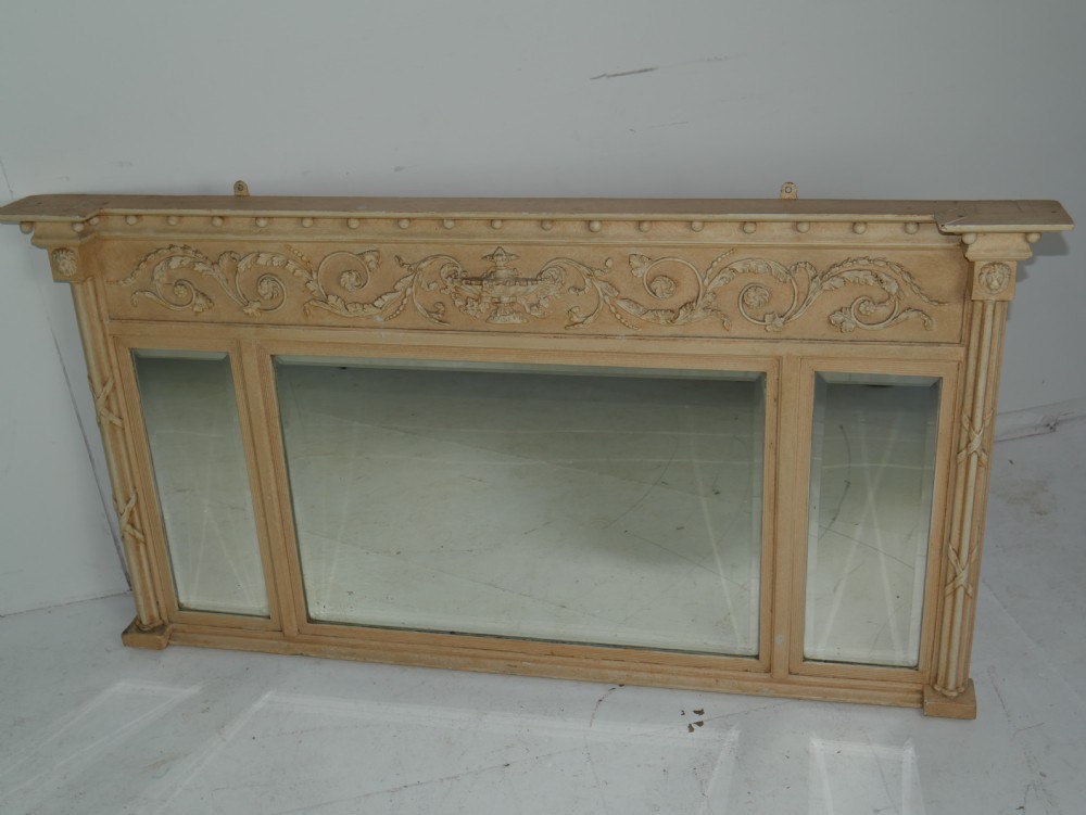 A wide cream-painted neoclassical overmantel mirror featuring three glass panels, an ornate carved urn frieze, and side pilasters. View X