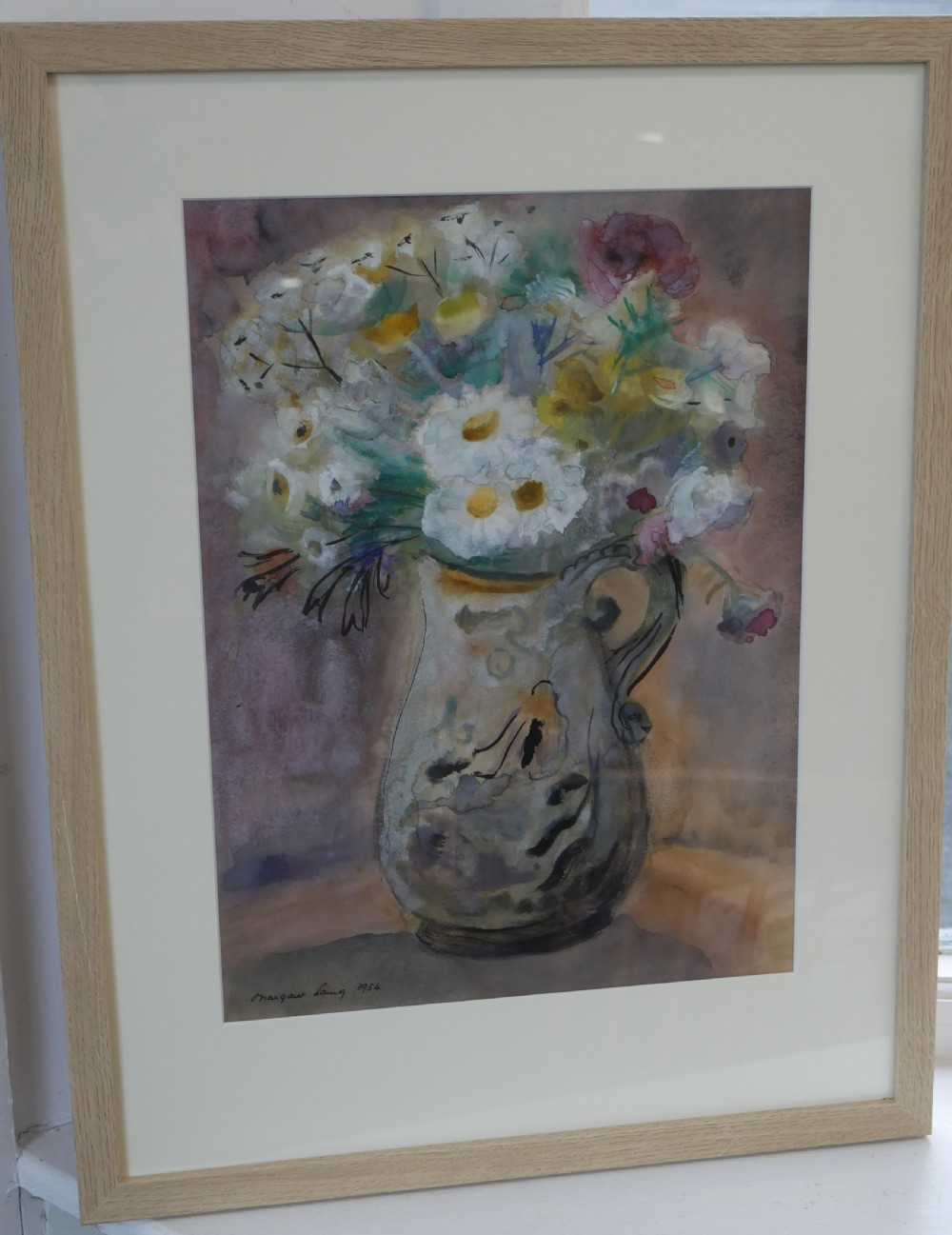 A framed watercolour painting depicting white and yellow flowers in a decorative cream jug, signed Margaret Laing and dated 1954.