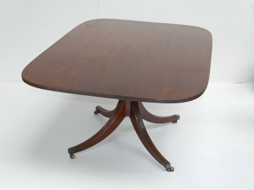 A Georgian mahogany centre table featuring a rounded rectangular top supported by a central pedestal with four reeded splayed legs and brass castors. View X