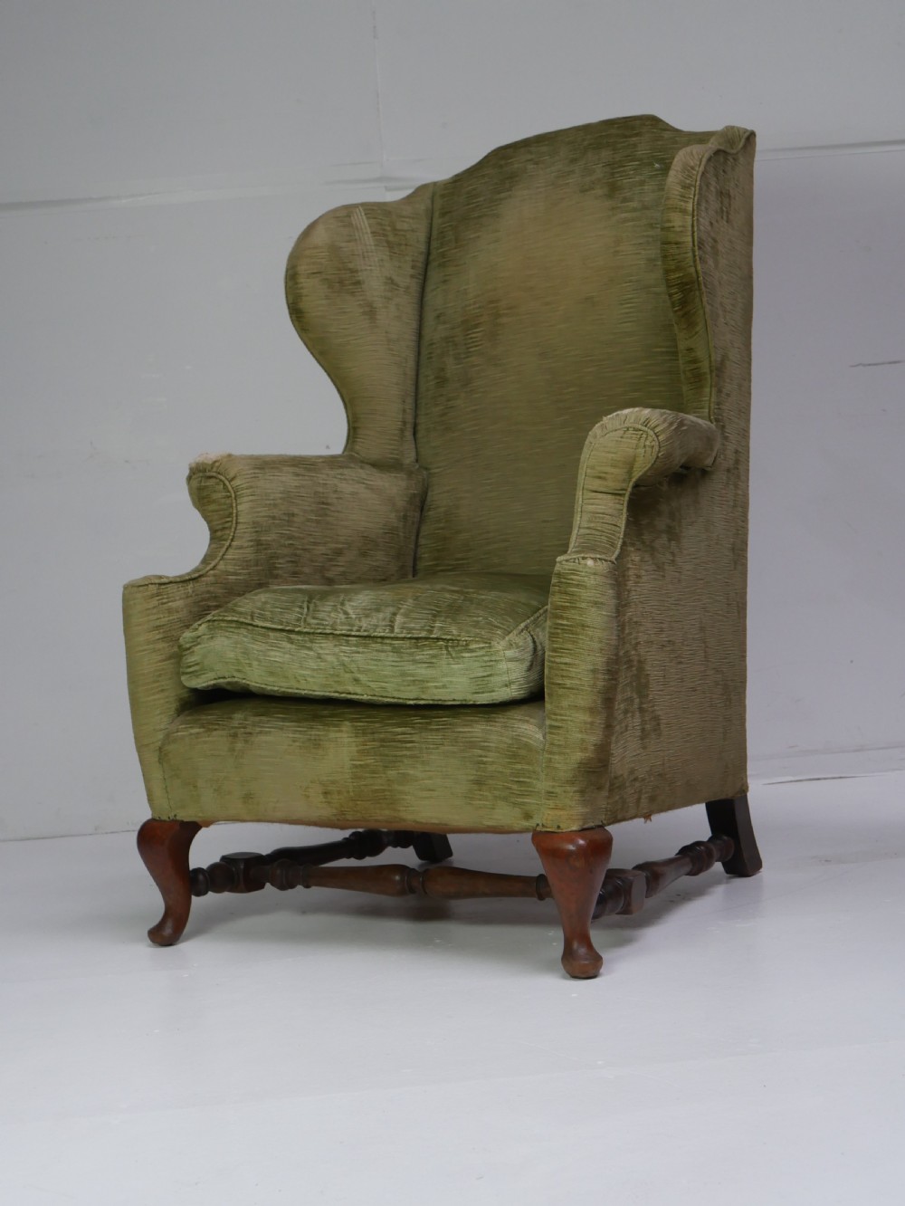 A high-backed wing armchair upholstered in textured olive green velvet with carved cabriole front legs and turned stretchers.