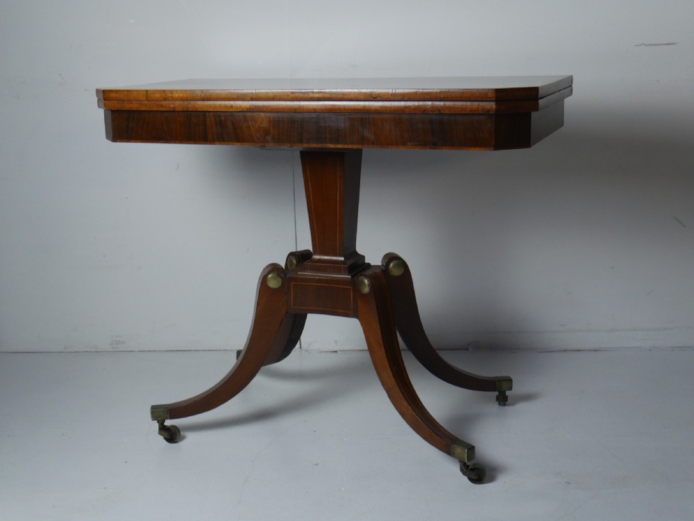 A Regency rosewood card table featuring a fold-over rectangular top above a tapered central pedestal and four splayed legs with brass castors.