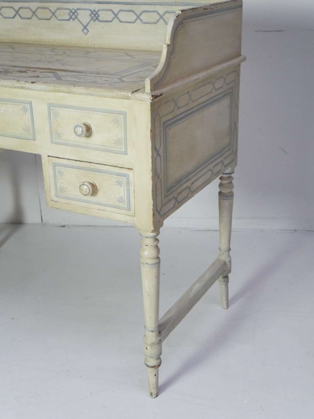 A 19th-century cream-painted wooden writing desk featuring five drawers, turned legs with stretchers, and a low gallery back decorated with light blue geometric painted borders. View II