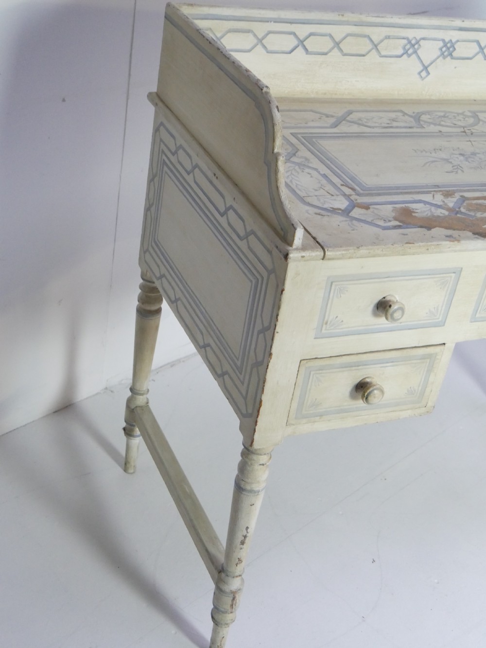 A 19th-century cream-painted wooden writing desk featuring five drawers, turned legs with stretchers, and a low gallery back decorated with light blue geometric painted borders. View XII