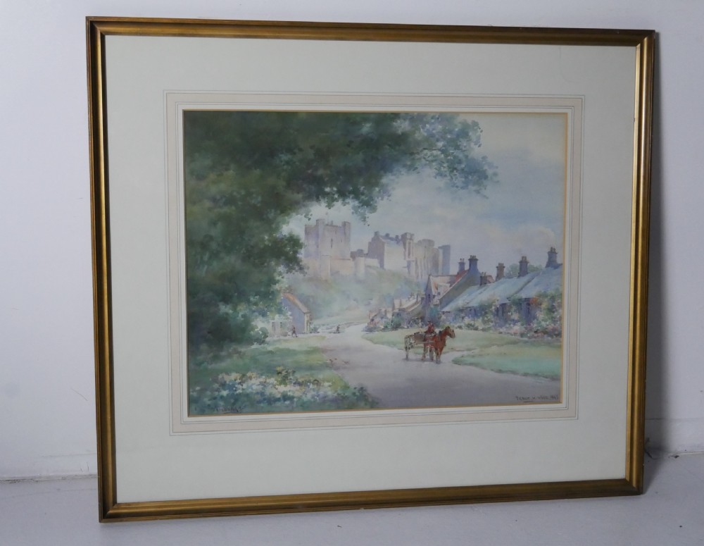 A framed watercolour painting depicting a village street with a horse and cart leading towards Bamburgh Castle under a soft sky.