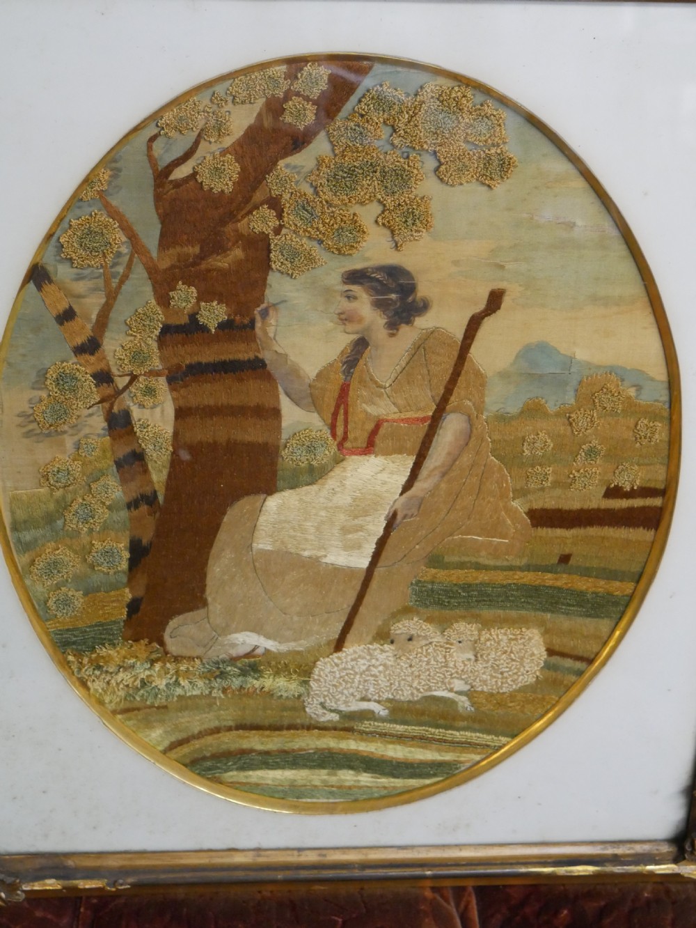 An early nineteenth-century oval crewel work embroidery depicting a shepherdess under a tree with sheep, set in a carved giltwood frame. View V