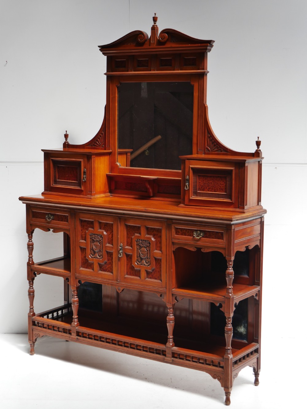 A late Victorian walnut and amboyna mirrored-back sideboard featuring carved panel doors, open tier shelving, turned spindles, and top finials.