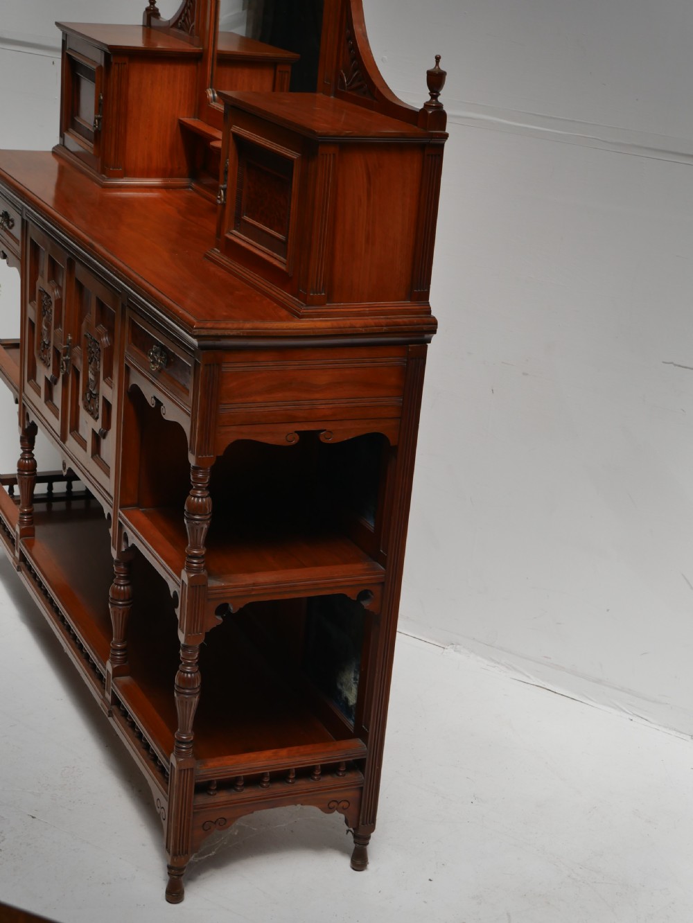 A late Victorian walnut and amboyna mirrored-back sideboard featuring carved panel doors, open tier shelving, turned spindles, and top finials. View XIII