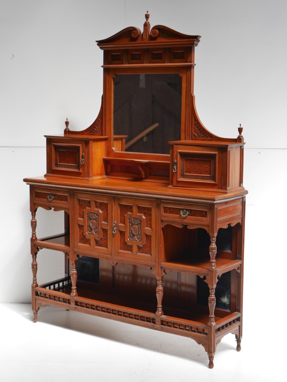 A late Victorian walnut and amboyna mirrored-back sideboard featuring carved panel doors, open tier shelving, turned spindles, and top finials. View XV