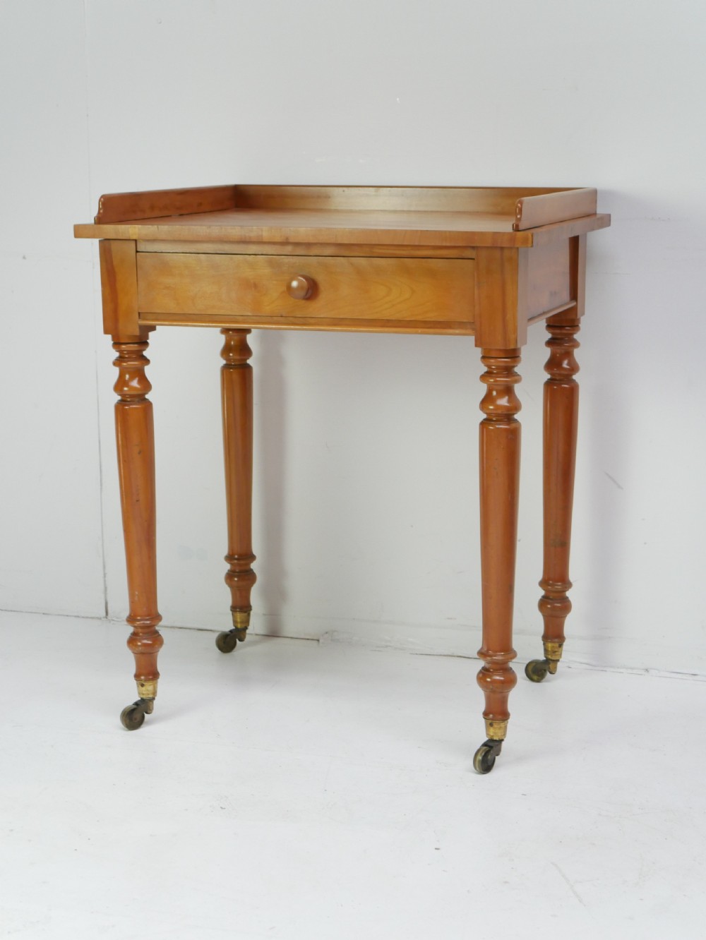 An antique satinbirch side table featuring a three-sided gallery top, single central drawer with a knob handle, turned legs, and brass castors.