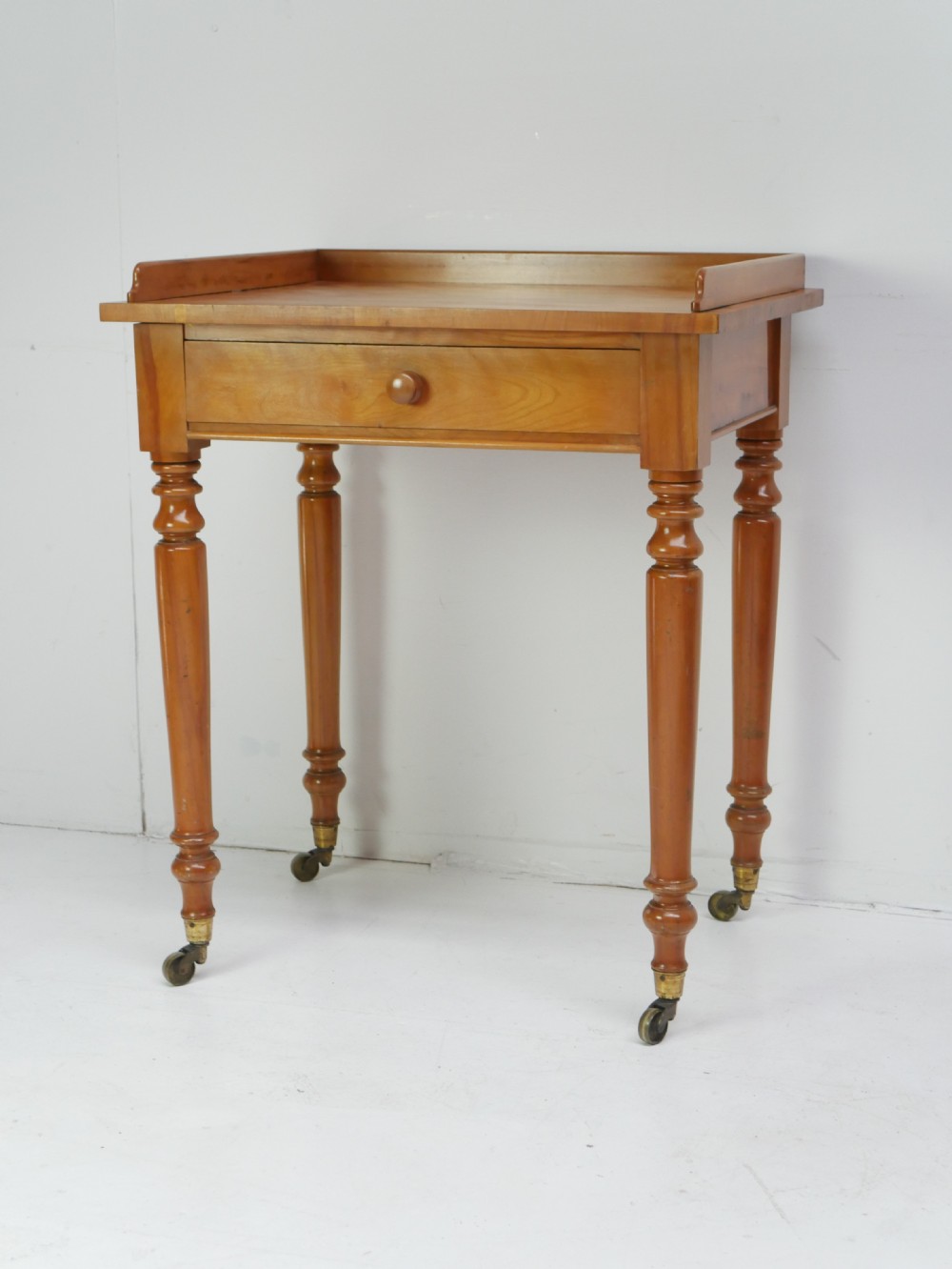 An antique satinbirch side table featuring a three-sided gallery top, single central drawer with a knob handle, turned legs, and brass castors. View XI