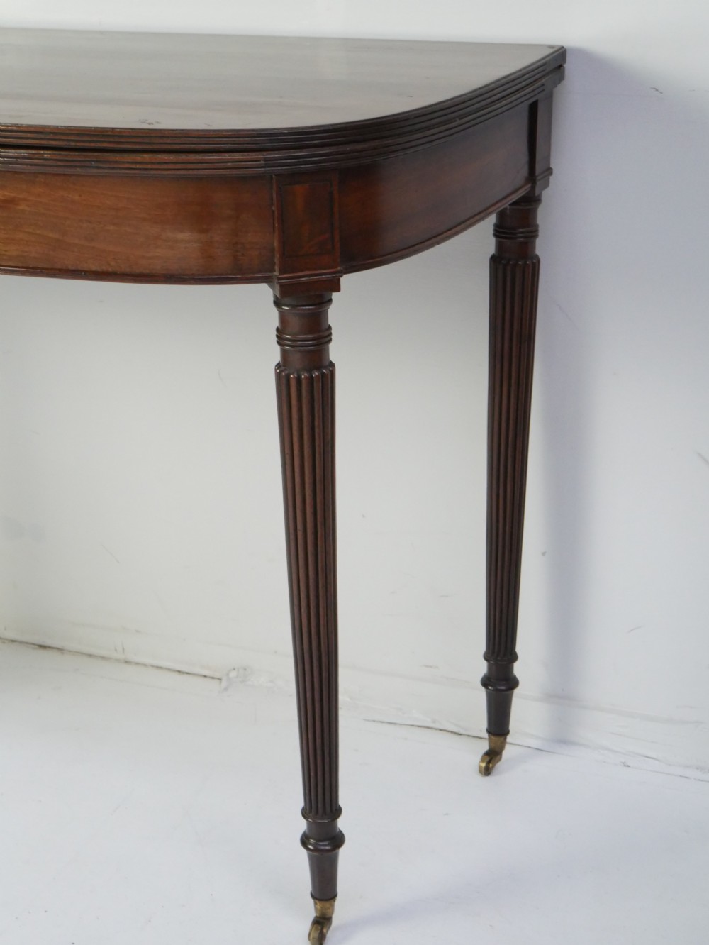 A Regency mahogany fold-over table featuring a demilune top, reeded legs, and brass castors. View II