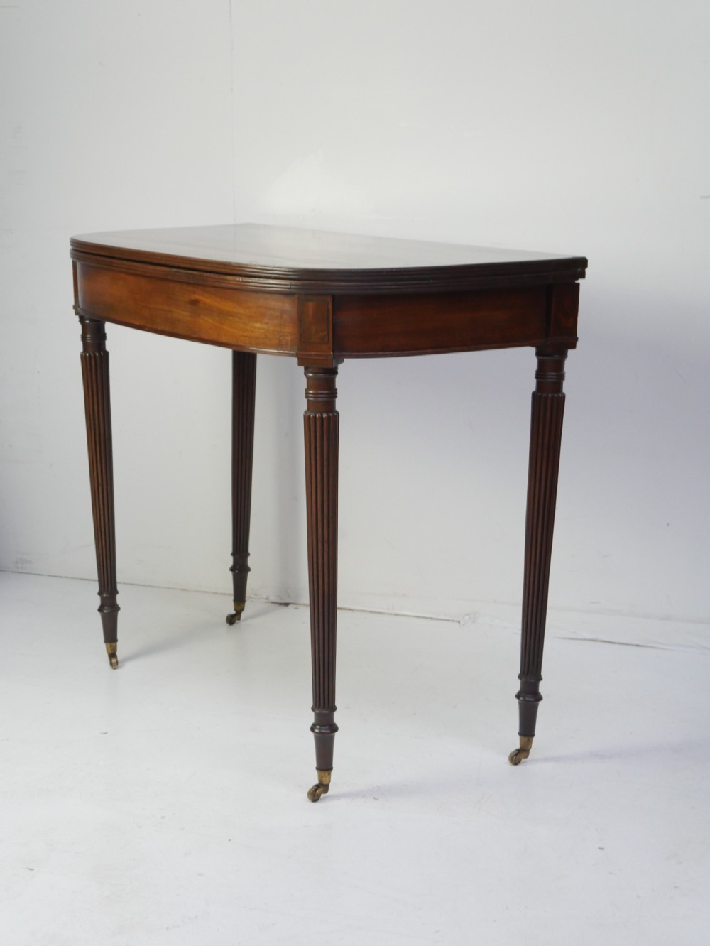 A Regency mahogany fold-over table featuring a demilune top, reeded legs, and brass castors. View V