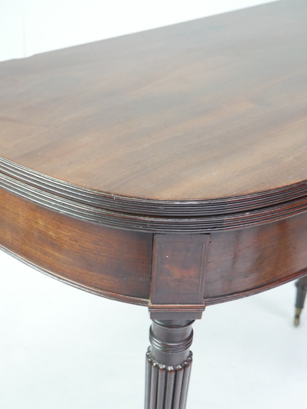 A Regency mahogany fold-over table featuring a demilune top, reeded legs, and brass castors. View X