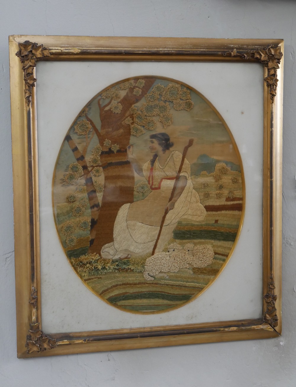 An early nineteenth-century oval crewel work embroidery depicting a shepherdess under a tree with sheep, set in a carved giltwood frame.