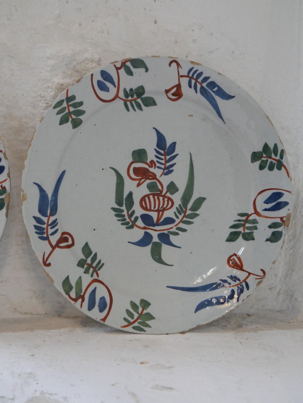 A pair of circular tin-glazed earthenware plates with off-white glaze, decorated with hand-painted polychrome floral motifs in blue, green, and iron-red. View VI