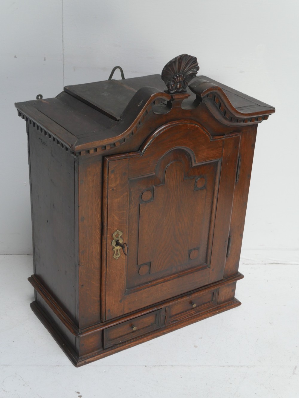 An 18th-century dark oak hanging cupboard featuring a carved shell cresting, a single fielded panel door with brass key escutcheon, and two small base drawers. View II