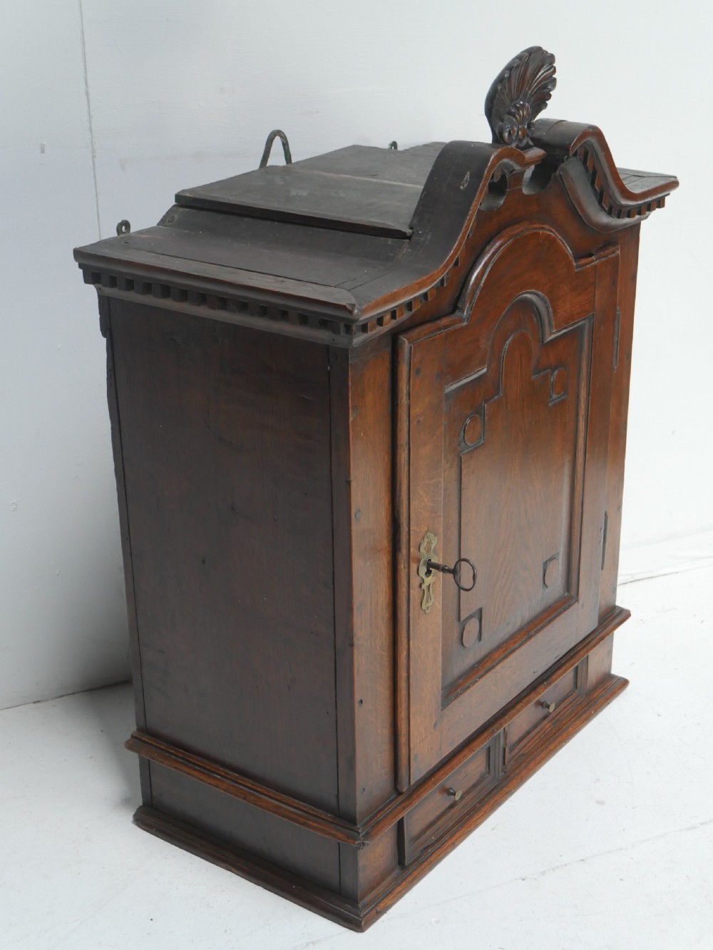An 18th-century dark oak hanging cupboard featuring a carved shell cresting, a single fielded panel door with brass key escutcheon, and two small base drawers. View III