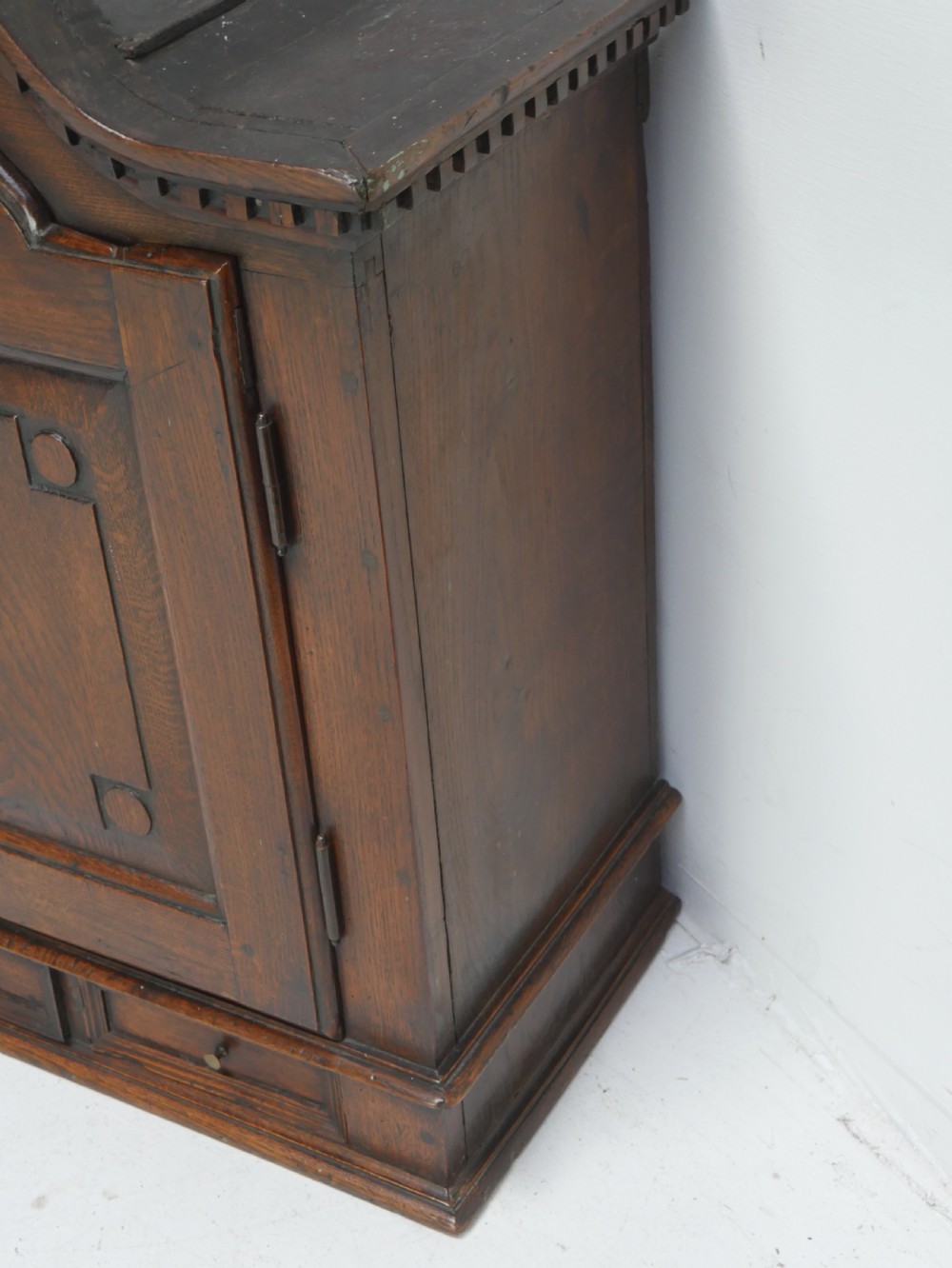 An 18th-century dark oak hanging cupboard featuring a carved shell cresting, a single fielded panel door with brass key escutcheon, and two small base drawers. View IV