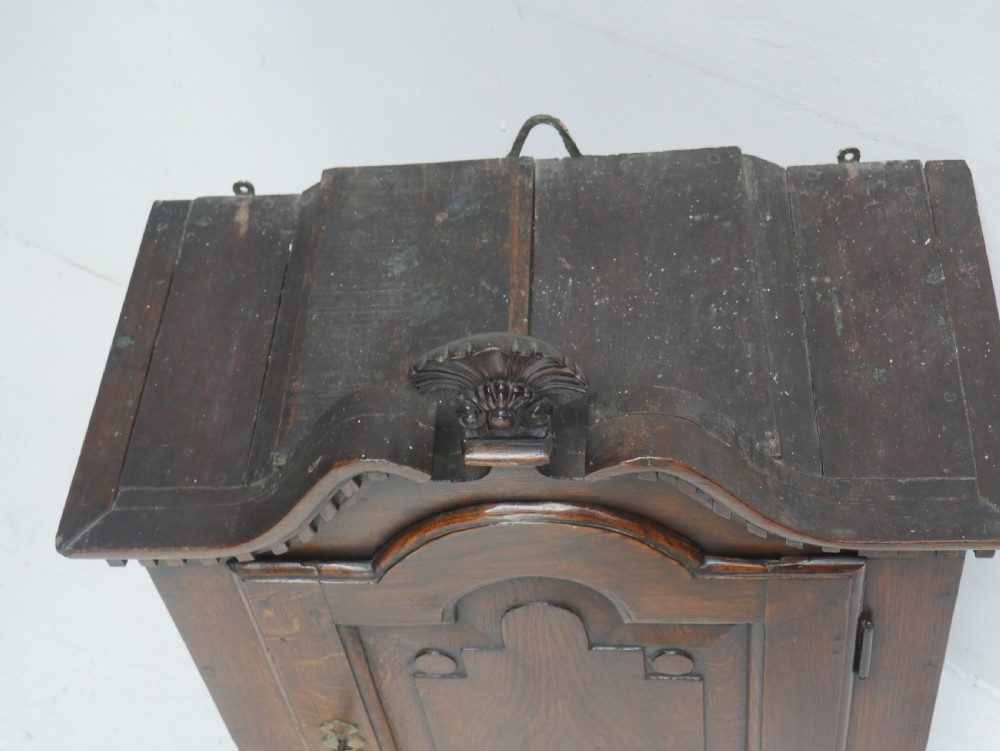 An 18th-century dark oak hanging cupboard featuring a carved shell cresting, a single fielded panel door with brass key escutcheon, and two small base drawers. View V