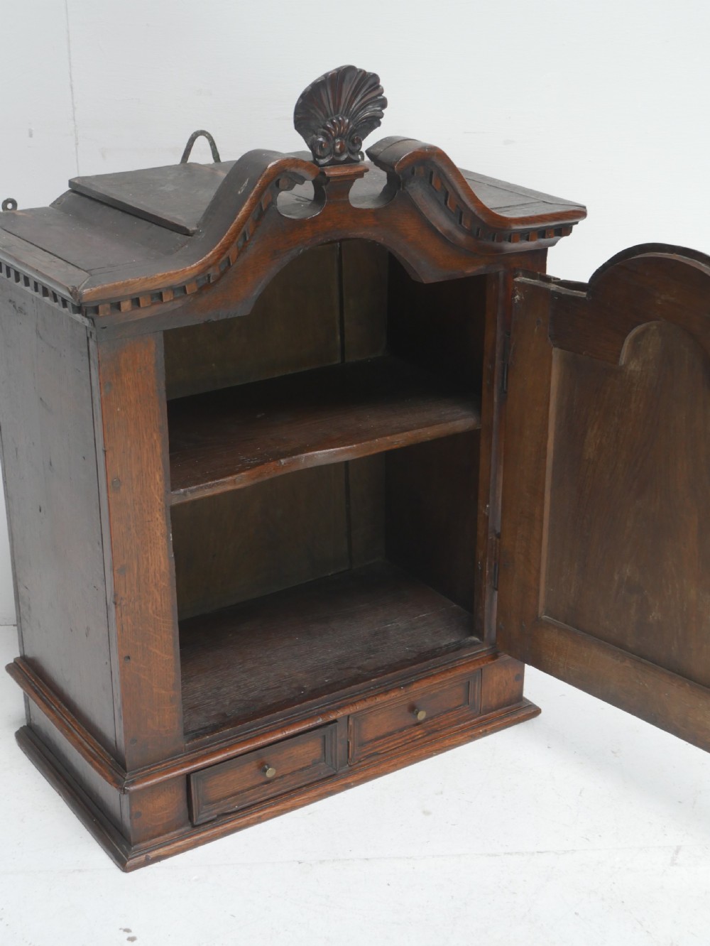 An 18th-century dark oak hanging cupboard featuring a carved shell cresting, a single fielded panel door with brass key escutcheon, and two small base drawers. View VII