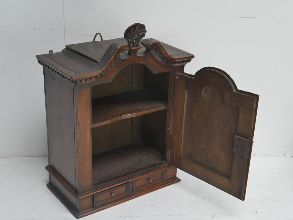 An 18th-century dark oak hanging cupboard featuring a carved shell cresting, a single fielded panel door with brass key escutcheon, and two small base drawers. View VIII