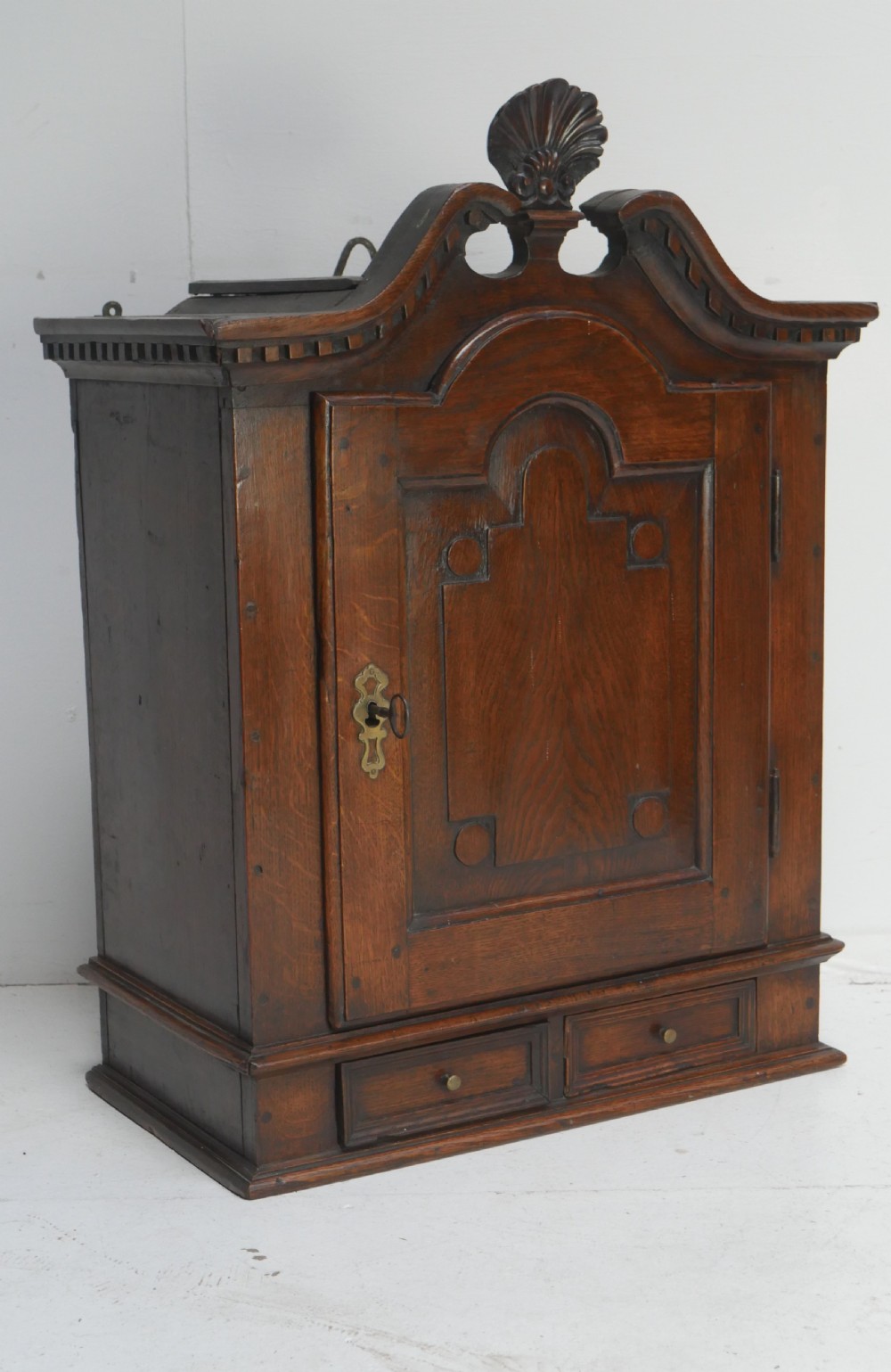 An 18th-century dark oak hanging cupboard featuring a carved shell cresting, a single fielded panel door with brass key escutcheon, and two small base drawers. View XV