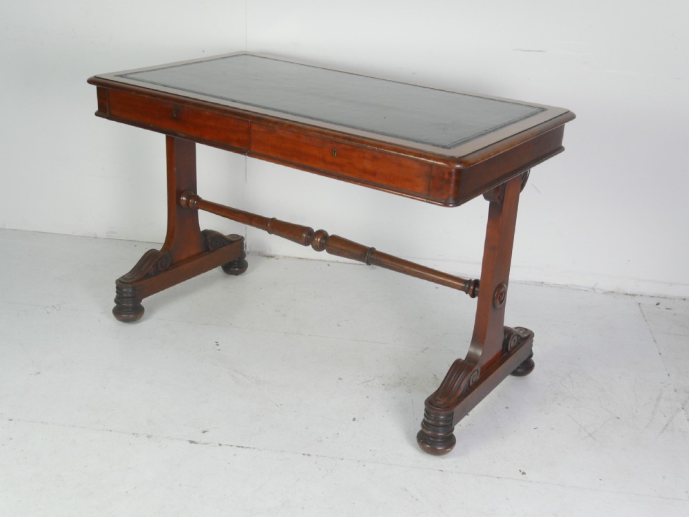 A mahogany writing desk featuring a dark leather inset top, two frieze drawers, and a turned stretcher connecting carved trestle end supports.