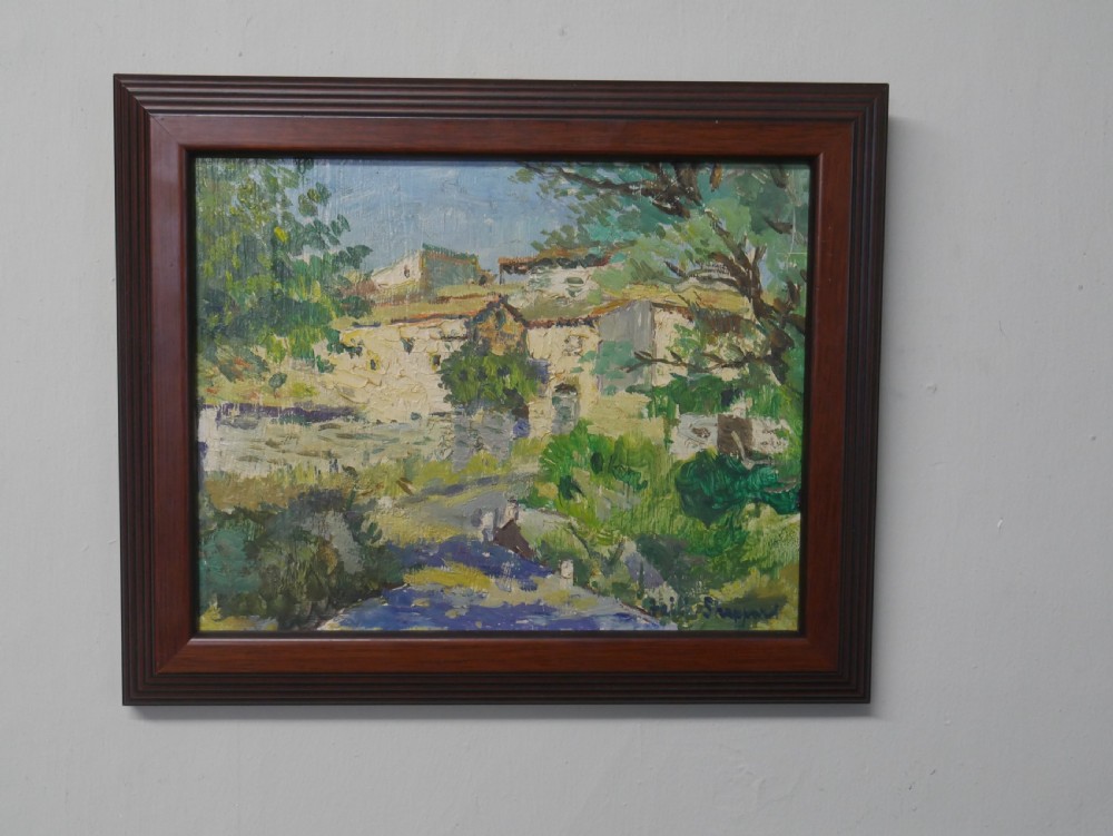 An impressionist oil painting depicting a sunny French village landscape with stone buildings and trees, presented in a dark wooden frame.