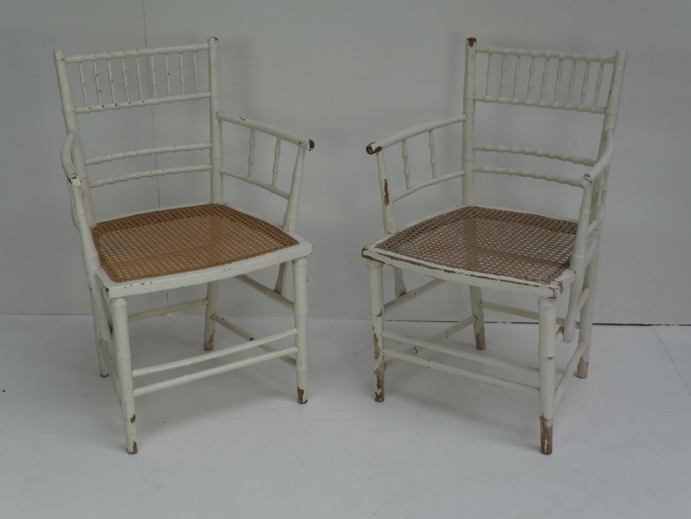 A pair of white painted wooden armchairs featuring turned spindle backrests, swept armrests, and woven cane seats.