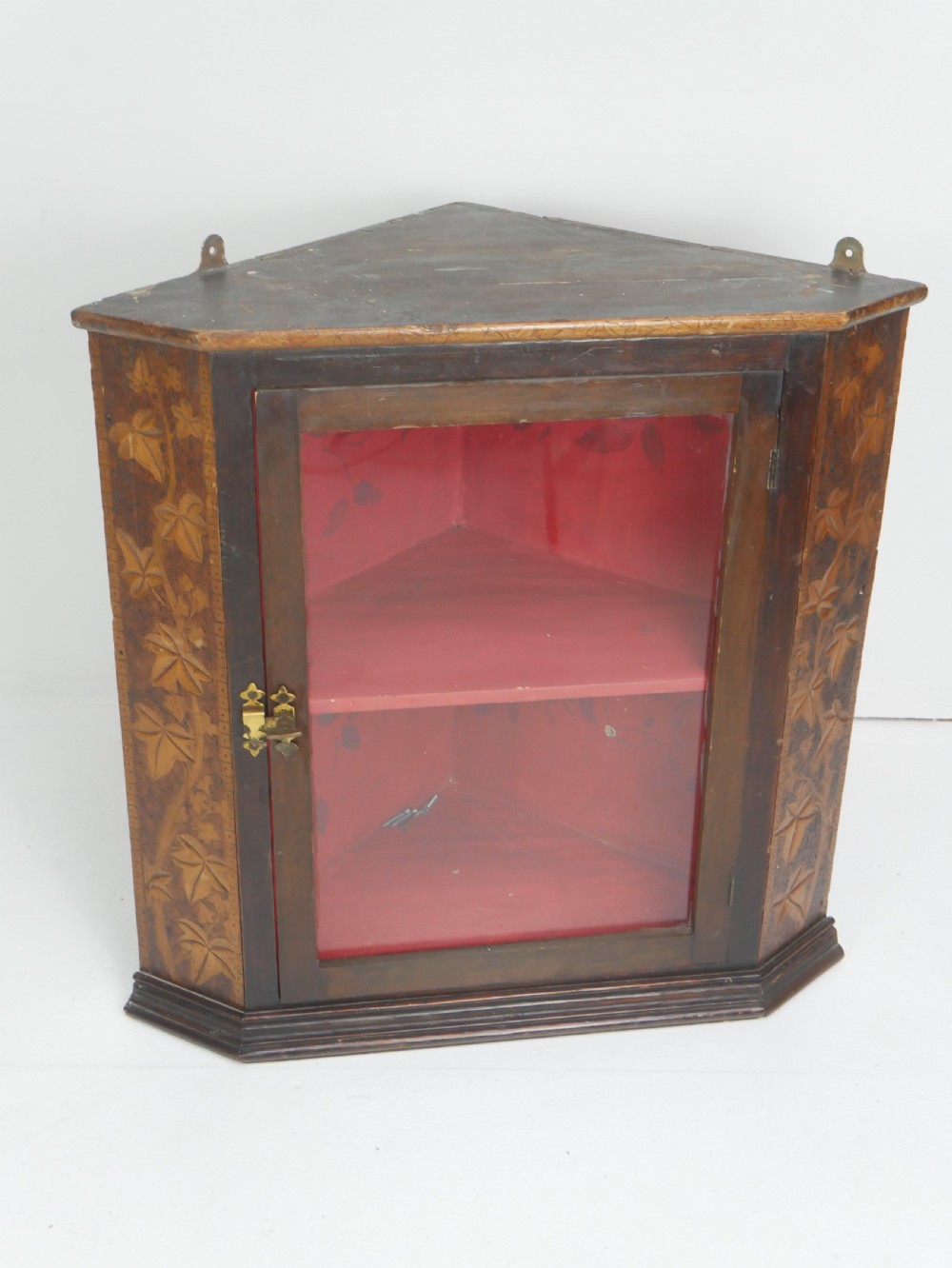 A dark carved wood wall-hanging corner cabinet featuring relief-carved ivy leaves on the side panels, a single glass door, a brass catch, and a red interior.