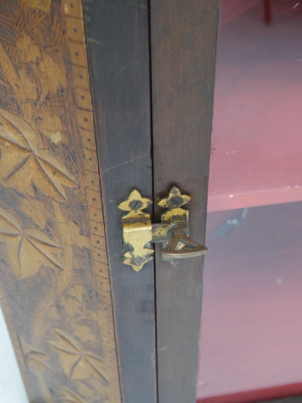 A dark carved wood wall-hanging corner cabinet featuring relief-carved ivy leaves on the side panels, a single glass door, a brass catch, and a red interior. View III