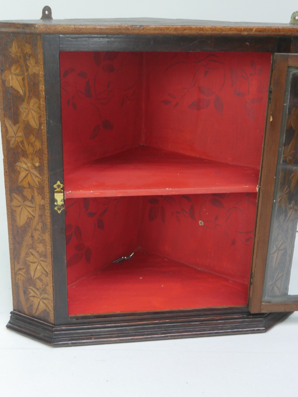 A dark carved wood wall-hanging corner cabinet featuring relief-carved ivy leaves on the side panels, a single glass door, a brass catch, and a red interior. View V