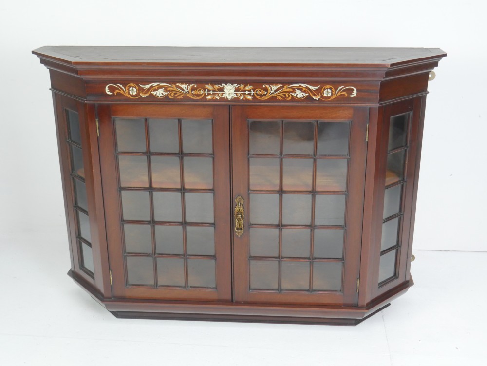 A wall-mounted mahogany display cabinet featuring glazed doors, canted side panels, decorative wood marquetry on the upper frieze, and a brass key escutcheon.