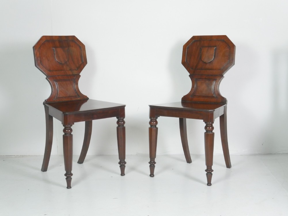 A pair of antique Regency mahogany hall chairs featuring octagonal carved backs with central shield motifs, solid timber seats, and turned front legs.