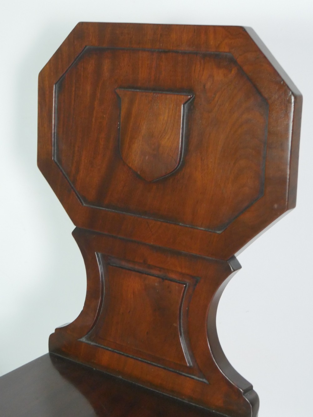 A pair of antique Regency mahogany hall chairs featuring octagonal carved backs with central shield motifs, solid timber seats, and turned front legs. View II
