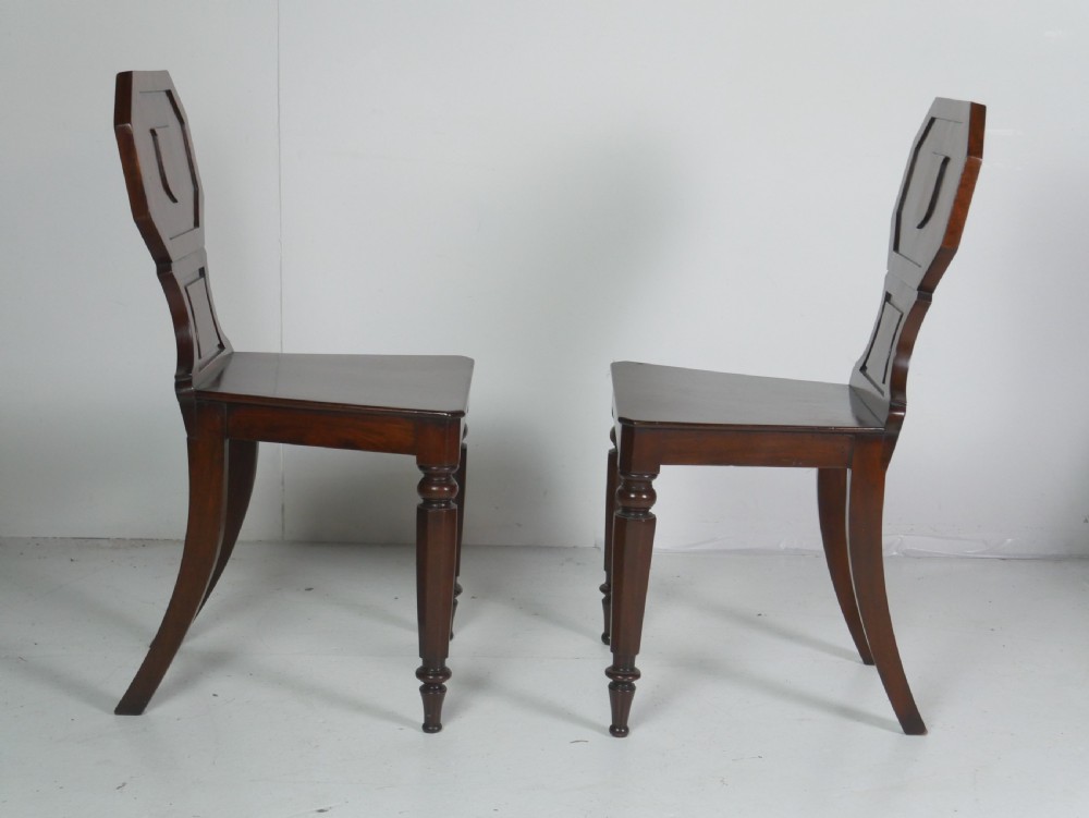 A pair of antique Regency mahogany hall chairs featuring octagonal carved backs with central shield motifs, solid timber seats, and turned front legs. View V